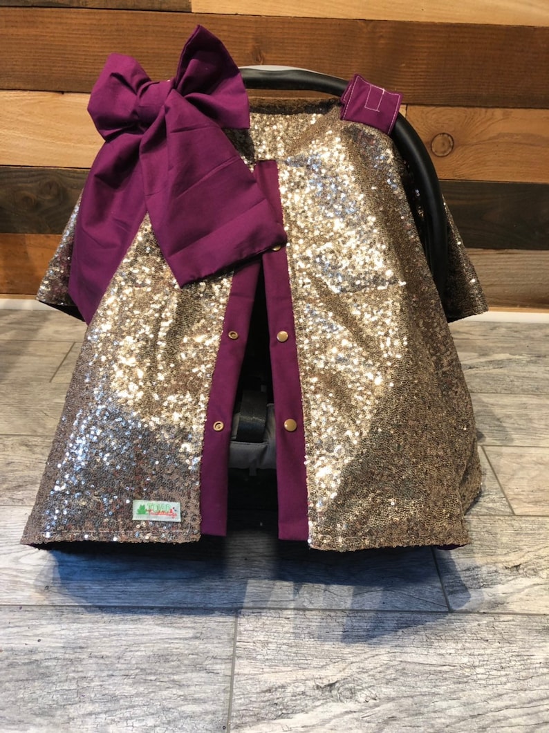 May include: Gold sequin car seat cover with a purple bow and buttons. The cover has a split down the back for easy access to the car seat straps.