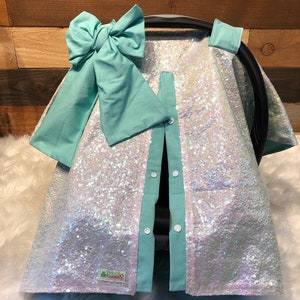 May include: A white iridescent sequin car seat cover with a light blue trim and a large light blue bow. The cover has a zipper down the front and snaps on the sides. The cover has a small label that says "Twinkle Covers".