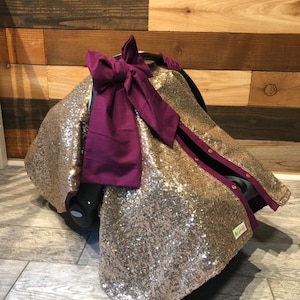 May include: Gold sequin car seat cover with a purple bow. The cover has a purple trim and snaps along the sides.