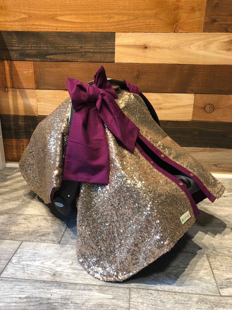 May include: Gold sequin car seat cover with a purple bow. The cover has a purple trim and snaps along the sides.