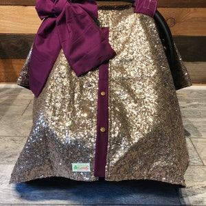 May include: A gold sequined car seat cover with a purple bow and a purple trim. The cover has a small green tag with the text "Green Frog Creations" on it.