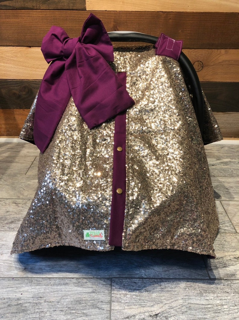 May include: A gold sequined car seat cover with a purple bow and a purple trim. The cover has a small green tag with the text "Green Frog Creations" on it.