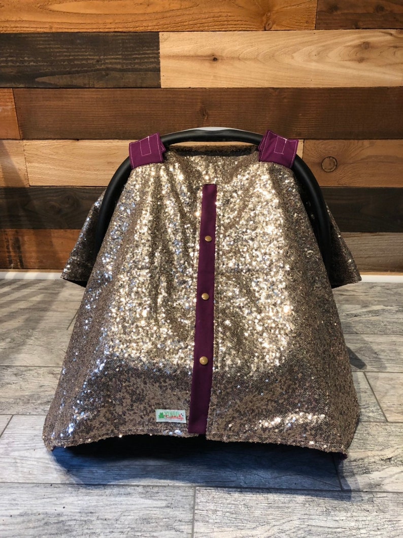 May include: A gold sequin car seat cover with purple trim and gold buttons. The cover has a small green tag with the text "Little Lambs" on it.