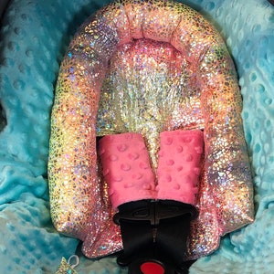 May include: A pink and iridescent car seat insert with a padded headrest and shoulder straps. The insert is designed to provide comfort and support for a baby in a car seat.