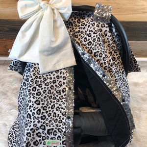 May include: A leopard print car seat cover with a white bow and silver sequins. The cover has a black lining and a small green tag with the word "TWINZ" on it.