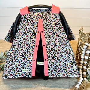May include: A gray and pink car seat cover with a colorful leopard print. The cover has a button closure and is made of soft fabric.