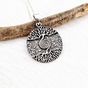 May include: A silver pendant necklace featuring a tree of life design with a spiral in the center. The pendant is round and has a textured surface.