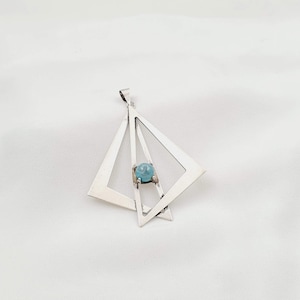 May include: A silver pendant featuring two overlapping geometric triangle frames with a single, round, light blue gemstone set in the center. The pendant includes a small silver bail at the top for attachment to a chain or cord.