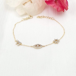 Könnte beinhalten: Ein goldenes Kettenarmband mit einem kleinen bösen Augen-Anhänger, einem Davidstern-Anhänger und einem kleinen Kreuz-Anhänger. Die Anhänger sind alle mit klaren Zirkonia-Steinen besetzt.