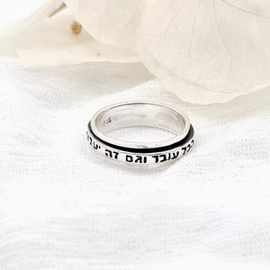 Puede incluir: Un anillo giratorio de plata con texto hebreo inscrito alrededor de la banda. El anillo está sobre un fondo de tela blanca.