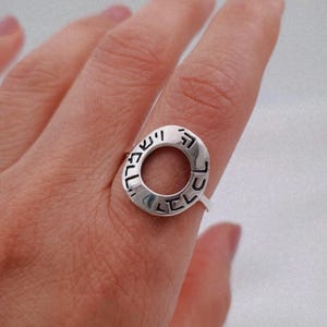 May include: A silver ring with an oval design. The ring features Hebrew text around the outer edge. The ring is worn on a finger.