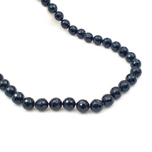 Puede incluir: Un collar de cuentas negras con cuentas redondas y facetadas. Las cuentas están ensartadas, creando una línea curva sobre un fondo blanco. El collar es una joya sencilla y elegante.