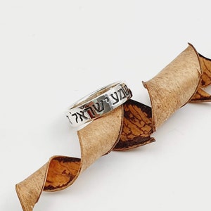 Puede incluir: Un anillo de plata con letras hebreas que dicen "Shema Yisrael" sobre un fondo marr&oacute;n.