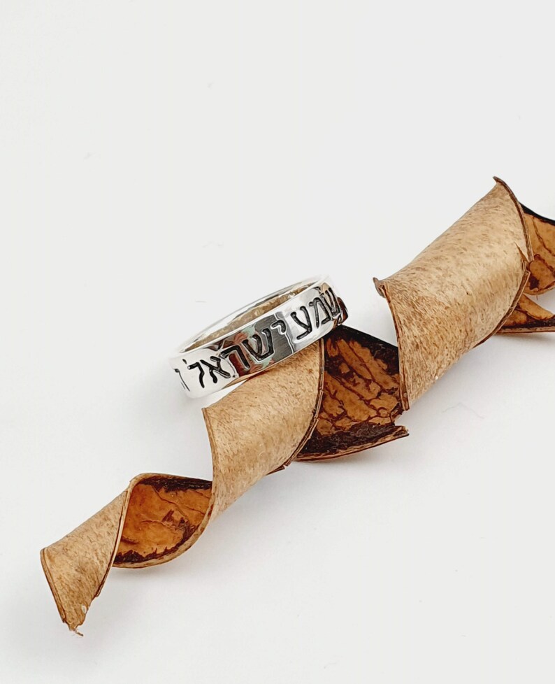 Puede incluir: Un anillo de plata con letras hebreas que dicen "Shema Yisrael" sobre un fondo marr&oacute;n.