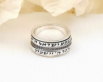 May include: A silver spinner ring featuring two bands engraved with Hebrew text in black lettering. Between the two engraved bands is a thin, textured silver band with a repeating pattern. The ring has a polished finish and a wide band design.