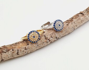 May include: Two silver and gold rings with blue and white evil eye designs. The rings are on a piece of driftwood.