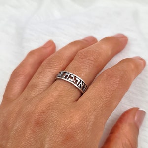 May include: A silver ring with Hebrew lettering. The ring is worn on a person's finger.