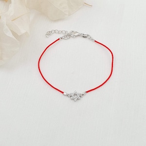 May include: A red string bracelet with a silver-colored star-shaped charm. The charm is encrusted with small, clear gemstones. The bracelet has a clasp closure and is displayed on a white surface.