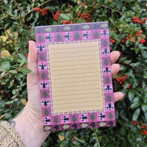 May include: A lined notepad with a decorative border featuring stylized birds, trees, and leaves in shades of pink, purple, and green. The notepad has a tan interior with horizontal lines for writing. The design is whimsical and nature-inspired.