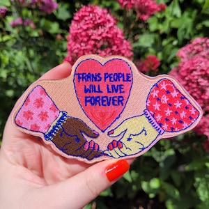 Puede incluir: Parche bordado rosa y rojo con dos manos sosteniendo un corazón rojo que dice "Trans People Will Live Forever".
