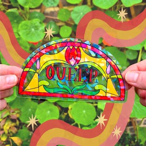 Op de afbeelding: Een kleurrijke sticker met het woord "QUEER" in vette letters, ontworpen in een glas-in-loodstijl. De sticker heeft een regenboog en sterren op een achtergrond van groen gebladerte.