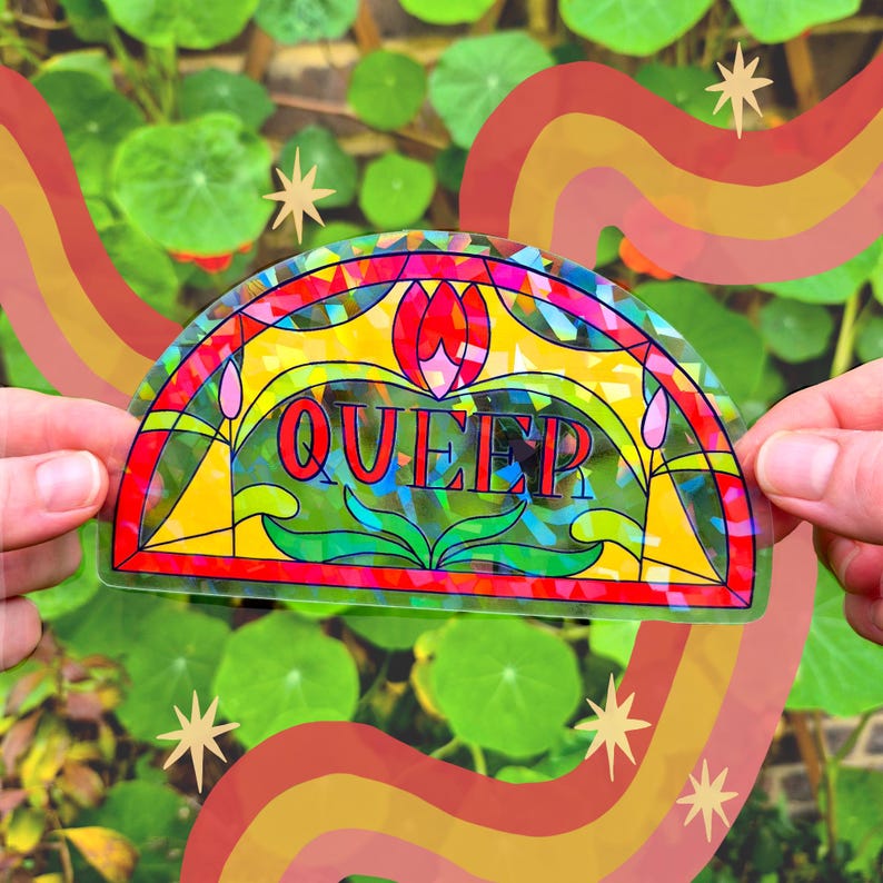 Op de afbeelding: Een kleurrijke sticker met het woord "QUEER" in vette letters, ontworpen in een glas-in-loodstijl. De sticker heeft een regenboog en sterren op een achtergrond van groen gebladerte.