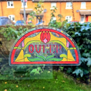 Op de afbeelding: Een kleurrijke, halfronde sticker met een glas-in-lood ontwerp. De sticker toont het woord "QUEER" in het midden, omringd door bloemen en geometrische patronen in rood, geel, groen en roze. De sticker is op een raam bevestigd.
