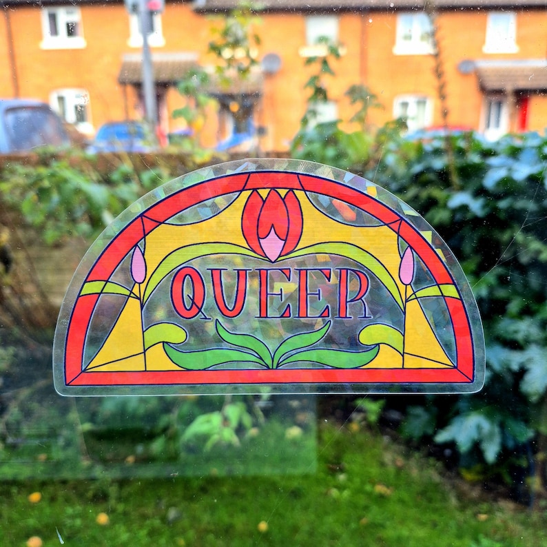 Op de afbeelding: Een kleurrijke, halfronde sticker met een glas-in-lood ontwerp. De sticker toont het woord "QUEER" in het midden, omringd door bloemen en geometrische patronen in rood, geel, groen en roze. De sticker is op een raam bevestigd.