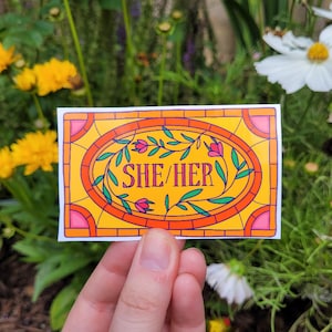 Puede incluir: Una pegatina amarilla y naranja con un diseño de vitral. La pegatina presenta un patrón floral y el texto "SHE/HER" en rosa.