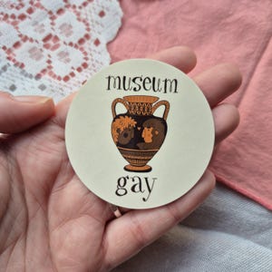 May include: A circular sticker with the words "museum gay" in black text. The sticker features a brown and black illustration of a vase with two figures inside. The sticker is held in a hand.