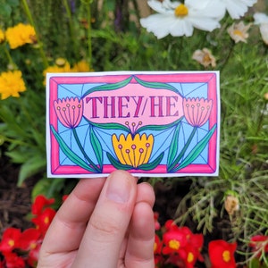 以下が含まれることがあります： ピンクと黄色の花と「THEY/HE」というピンクの文字が書かれたステンドグラスのデザインのステッカーです。