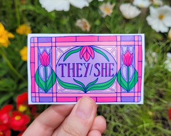 May include: A pink and purple sticker with a stained glass design featuring two pink tulips and the text "THEY/SHE".