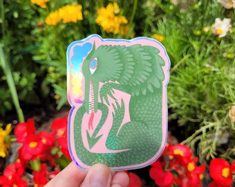 Könnte beinhalten: Ein holografischer Aufkleber mit einem grünen Drachen auf rosa Hintergrund. Der Drache hat einen langen Hals, ein grinsendes Maul und eine Regenbogenwolke. Der Aufkleber wird vor einem Hintergrund aus Blumen gehalten.