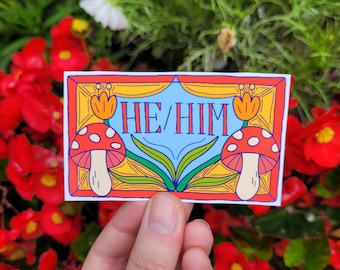 May include: A colorful sticker with a blue background and the text "HE/HIM" in white. The sticker features two red and white mushrooms with yellow stems and green leaves. The sticker is framed in a yellow and red border.
