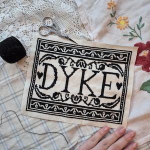May include: A cross-stitch with the word "DYKE" in black, surrounded by decorative borders. A black thread ball and small scissors are visible. The piece rests on patterned fabric, with a hand in the lower right.