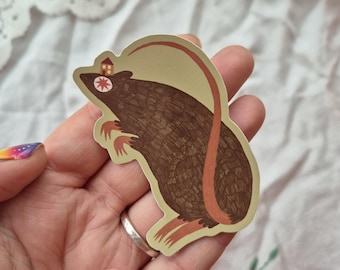 May include: A brown rat sticker with a house on its head and a long, curved tail. The rat has small orange feet and a red star for an eye. The sticker is on a light green background.