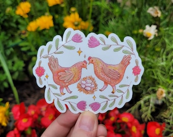 May include: A white sticker with a floral border and two pink chickens. The chickens are holding a nest of white eggs. The sticker is perfect for adding a touch of whimsy to any project.