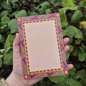 May include: A rectangular notepad with a pink center and a decorative border featuring running hares in brown, orange, and gray. The border is set against a green background with a pink and yellow trim. A star design is in each corner.