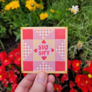 以下が含まれることがあります： ピンクと赤の継ぎ合わせのキルトブロックで、黄色い中心にピンクの文字で「Stay Soft」と書かれています。ブロックは黄色の縁取りで囲まれています。