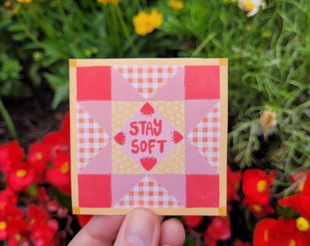 May include: A pink and red patchwork quilt block with a yellow center featuring the text "Stay Soft" in pink lettering. The block is surrounded by a yellow border.
