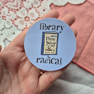 May include: A circular sticker in pale blue, featuring the words "library" and "radical" in black script. The sticker has an illustration of a book titled "The Picture of Dorian Gray" by Oscar Wilde.