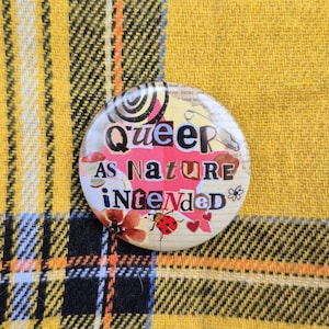 May include: A circular pinback button featuring a collage design with the text "Queer as Nature Intended" in mixed black and dark blue lettering. The background includes a pink star, a red ladybug, floral elements, and lined paper textures.