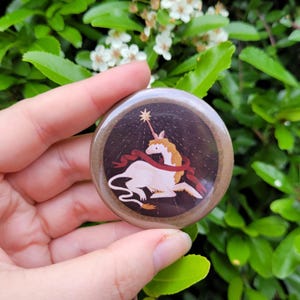 May include: A circular badge featuring a white unicorn with a golden mane and a red ribbon, set against a dark brown backdrop with a shining star. The badge has a brown border. The design is whimsical.