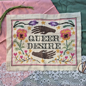 May include: A rectangular cross-stitch embroidery on off-white fabric featuring the text "QUEER DESIRE" in black letters. The design includes two dark brown hands, two large pink and yellow pansies, purple flowers, small yellow stars, and pink hearts.