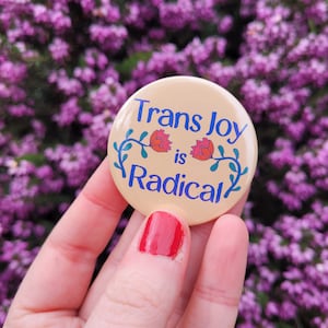 Puede incluir: Un botón beige con el texto "Trans Joy is Radical" en letras azules y dos flores rosas con tallos verdes. El botón está siendo sostenido por una mano contra un fondo de flores de brezo púrpura.