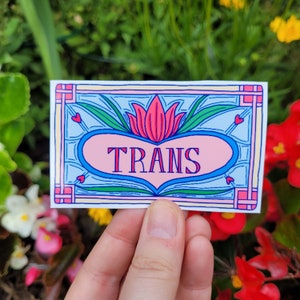 K&ouml;nnte beinhalten: Ein Aufkleber mit einem rosa Herz und einem blauen Hintergrund. Das Herz hat einen dekorativen Rand und das Wort "TRANS" ist in rosa Buchstaben geschrieben.