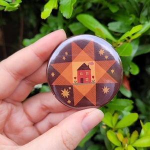 May include: A round button with a quilt-inspired design featuring a red house with a smoking chimney, surrounded by star and geometric patterns in shades of brown, orange, and yellow. The button is held in a hand against a backdrop of green foliage.