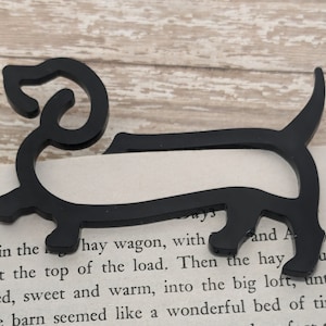 May include: Black metal dachshund bookmark. The bookmark is shaped like a dachshund dog and is sitting on a book with the text "barn in the hay wagon, with ing at the top of the load. Then the hay hoisted, sweet and warm, into the big loft, until the whole barn seemed like a wonderful bed of timothy 43 and A rid- uld be" visible.