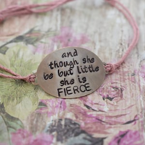 May include: A pink string bracelet with a silver charm that reads "and though she be but little she is fierce".