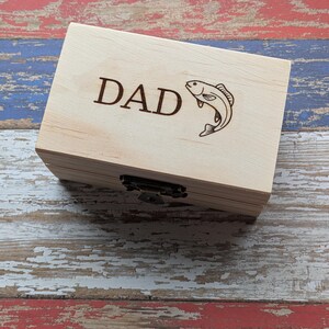 May include: A rectangular light-colored wooden box featuring a dark brown laser-engraved design on the lid. The engraving includes the word DAD in capital letters followed by a graphic illustration of a fish. The box has a metal latch closure.